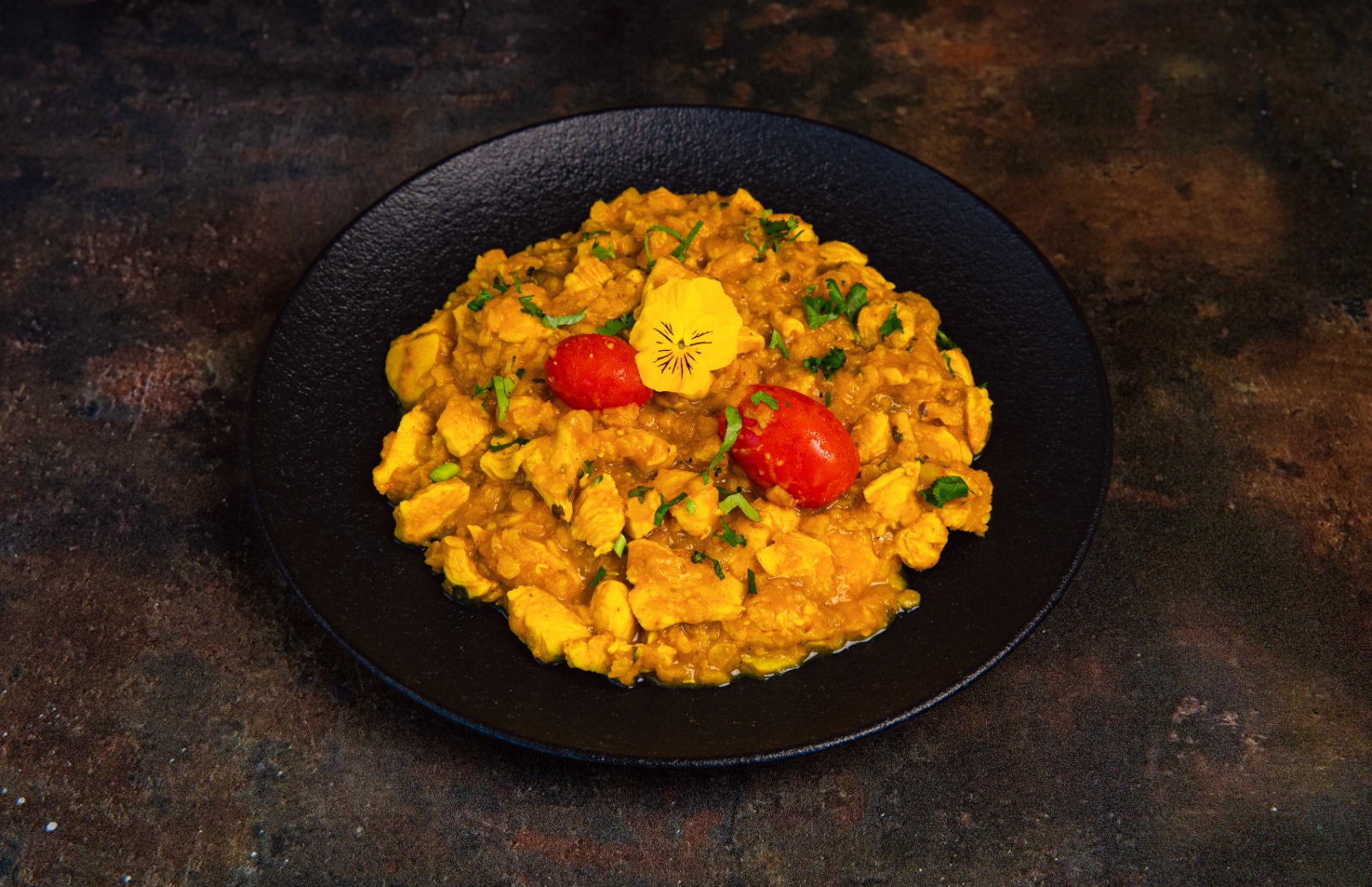 Dark plate with vibrantly coloured chicken curry garnished with herbs and a pansy